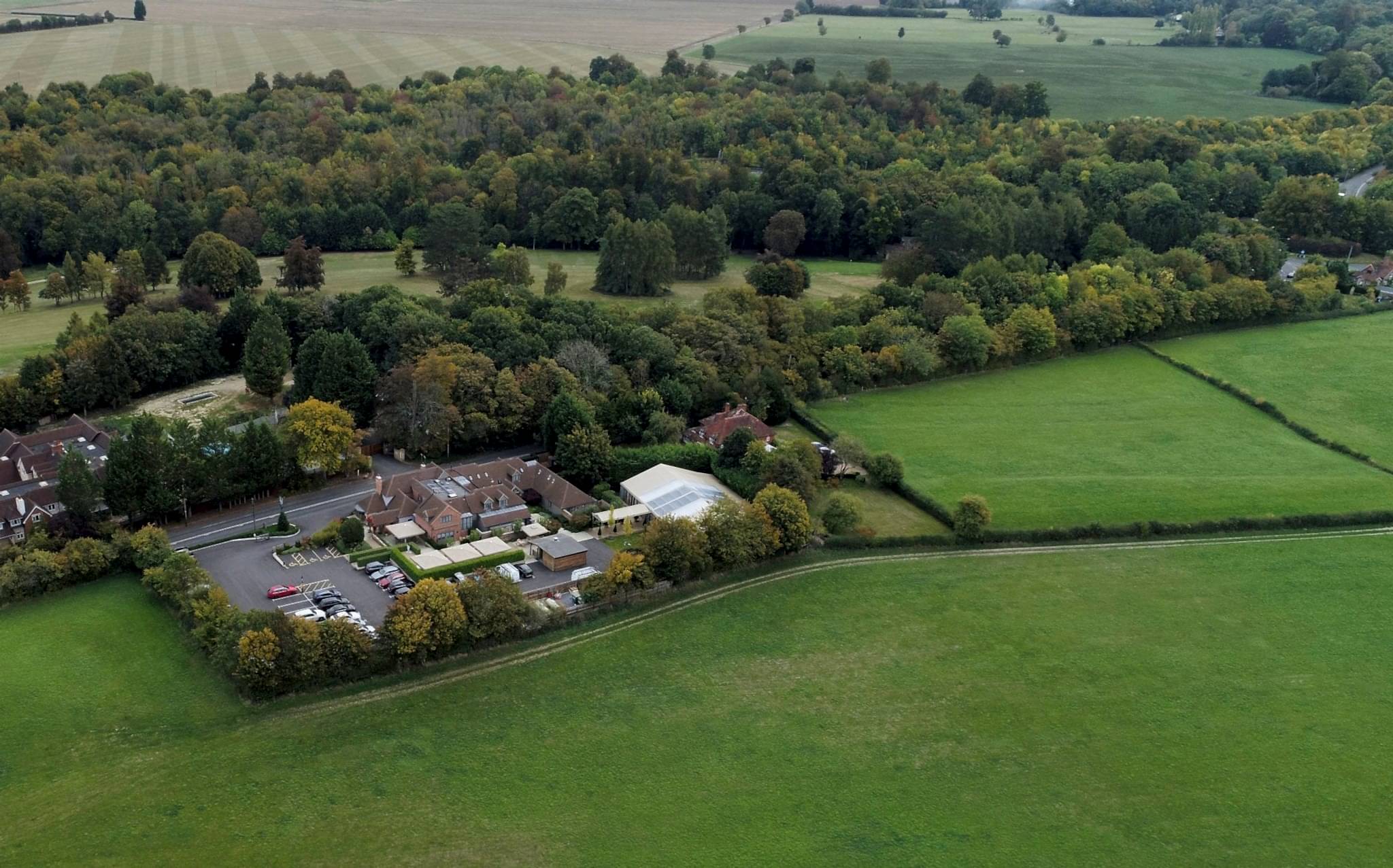 Hurley House - Aerial view of a rural landscape with a cluster of buildings surrounded by trees and open land. The area features fields and dense woodland, with a small parking lot adjacent to the buildings. Hills and more trees are visible in the background.