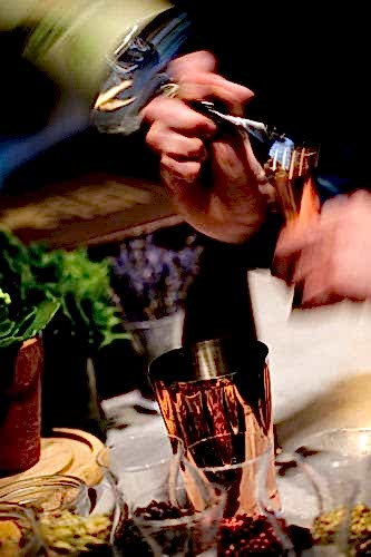 Hurley House - A person vigorously shakes a cocktail shaker surrounded by various ingredients and garnishes, including herbs and spices, on a countertop. The motion creates a blur, emphasizing the dynamic action.