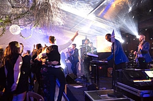 Hurley House - A lively party scene with a band performing on stage, featuring a keyboardist and guitarist in blue jackets. Attendees dance energetically, and the atmosphere is enhanced by bright lights, disco balls, and hanging decorations under a transparent tent roof.