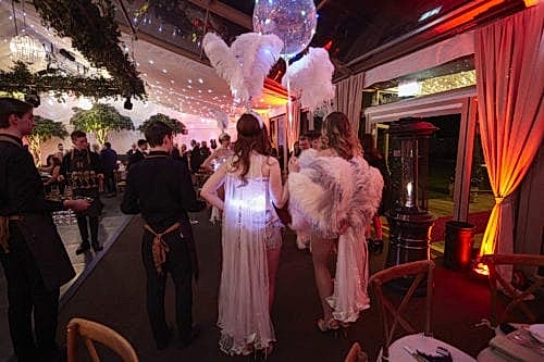 Hurley House - A glamorous indoor event with people in elegant dresses and feathered accessories. Attendees include servers carrying trays. The venue is adorned with lights, plants, and draped curtains. A festive atmosphere is evident.