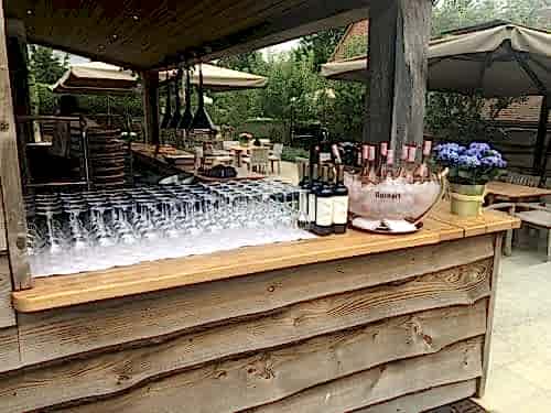 Hurley House - Outdoor bar setup featuring rows of wine glasses, bottles of red wine, and a large bowl of ice holding several bottles of rosé. Wooden bar counter under a shaded pavilion, with outdoor seating and greenery in the background.
