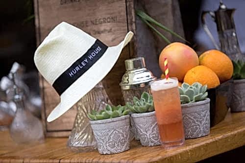 Hurley House - A straw hat with a Hurley House band sits on a wooden table next to a cocktail, succulents, and citrus fruits. A shaker and glass bottles are in the background.