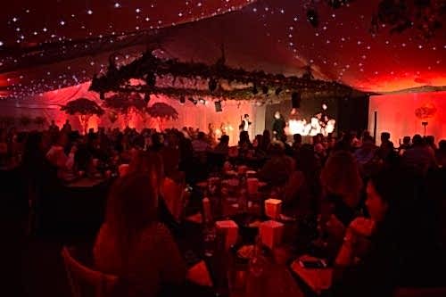 Hurley House - A lively indoor event with a red and white starry-themed ceiling. Attendees sit at tables watching performers on stage. The setting has a festive ambiance with dim lighting and decorative foliage hanging above.