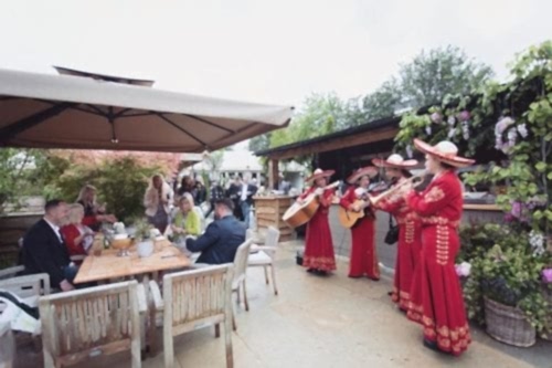 Hurley House - A mariachi band in red outfits performs outdoors for people sitting at wooden tables. The setting includes leafy green plants, umbrellas, and a relaxed atmosphere.