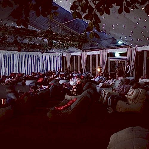 Hurley House - People are seated on large, cushioned recliners in a dimly lit room, watching a projection at the front. The ceiling is decorated with string lights and greenery, creating a cozy ambiance. The audience appears relaxed and engaged.