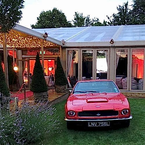 Hurley House - A vintage red sports car parked on grass in front of a clear-roofed event tent with warm lighting. Triangular topiary trees line a red carpet leading into the tent.