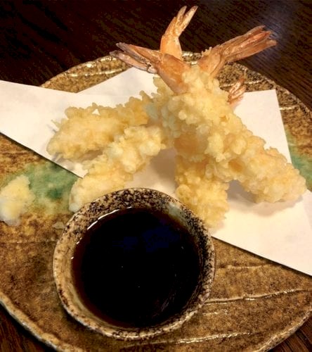 Hurley House - Three golden shrimp tempura pieces on a brown ceramic plate with a small bowl of soy sauce on the side. White paper is placed beneath the tempura.