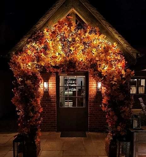 Hurley House - A small brick building with a fall-themed arch made of colorful autumn leaves and twinkling lights at the entrance. Two wall-mounted lanterns illuminate the scene, adding warmth to the cozy, night-time setting.