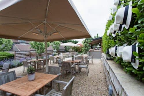Hurley House - Outdoor patio with wooden tables and chairs under large umbrellas. A wall adorned with white hats on the right adds a decorative touch. Lush greenery surrounds the area, creating a cozy and inviting atmosphere.