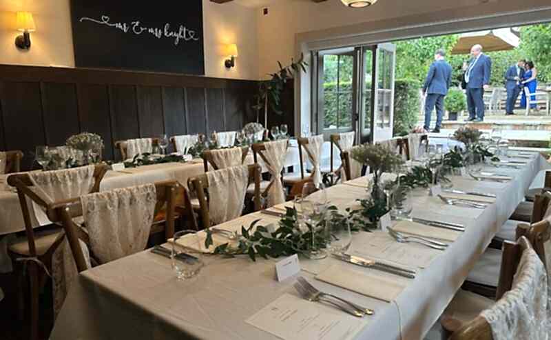 Hurley House - An elegantly set table with white linens and greenery is prepared for a luxury wedding reception in Berkshire. White chairs with lace decorations line the table, beneath a sign reading Mr & Mrs in cursive. Through open glass doors, two people talk outside.