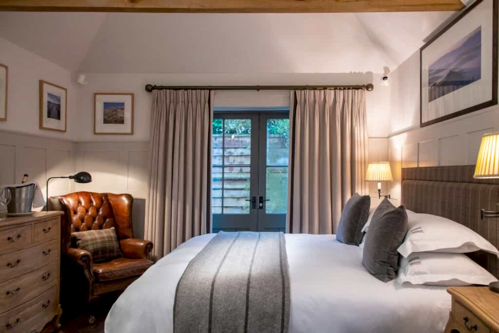 Hurley House - A cozy bedroom with a large bed covered in white linens and a gray throw, a brown leather armchair, wooden dresser, framed pictures on the walls, and double doors with curtains leading outside. Lamps provide warm lighting.