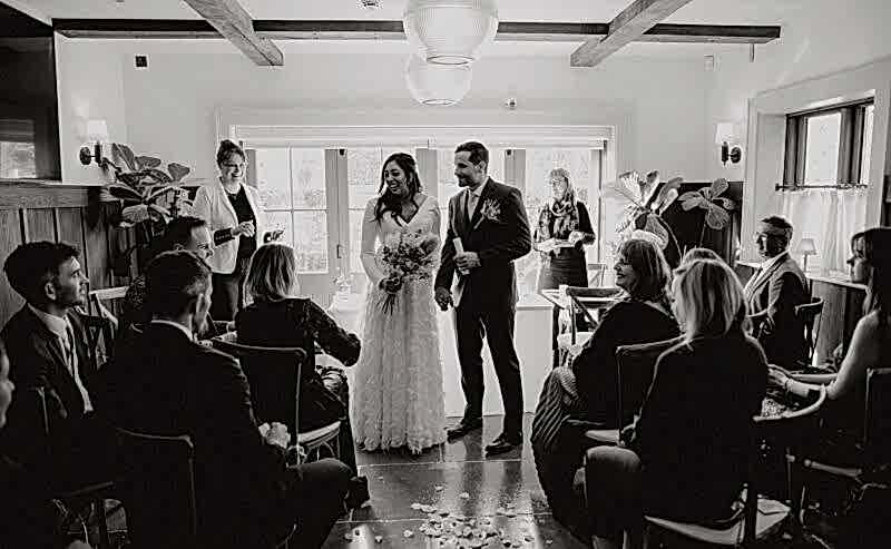 Hurley House - A black and white photo of a wedding ceremony. A bride and groom stand together, smiling, at the front of a room filled with seated guests. Natural light streams in from windows behind them, illuminating rose petals scattered on the floor.