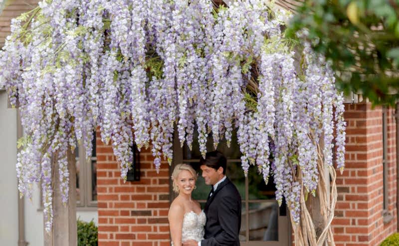 Hurley House - A bride and groom stand beneath a wooden archway adorned with cascading purple wisteria flowers, smiling and embracing outside a brick building surrounded by lush greenery.