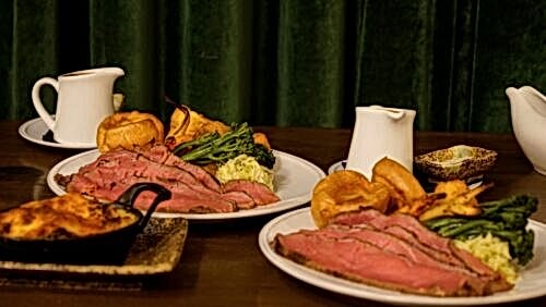 Hurley House - Plates of sliced roast beef with Yorkshire pudding, broccoli, potatoes, and gravy served on a wooden table, with sauce jugs and side dishes, in front of a dark green curtain.