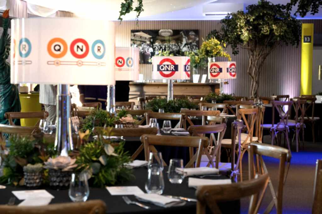 Hurley House - A decorated indoor dining area with round tables, wooden chairs, potted plants, and tall lamps featuring QNR logos. Table settings include glasses and cutlery; greenery and framed photos adorn the space.