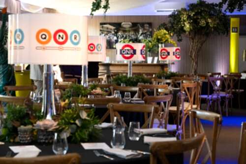 Hurley House - A decorated indoor dining area with round tables, wooden chairs, potted plants, and tall lamps featuring QNR logos. Table settings include glasses and cutlery; greenery and framed photos adorn the space.