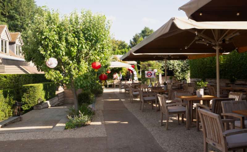Hurley House - Outdoor restaurant seating area with wooden tables and chairs under large umbrellas, surrounded by greenery and trees decorated with red lanterns on a sunny day. Pathway runs through the middle of the area.