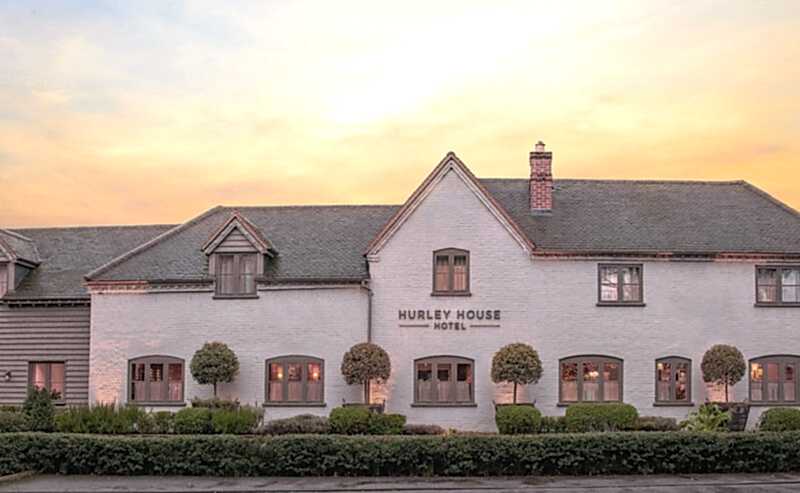 Hurley House - A charming hotel with a rustic exterior and neatly trimmed bushes, set against a peaceful sunset sky. The building is painted white with gray accents and has a sign that reads Hurley House Hotel.