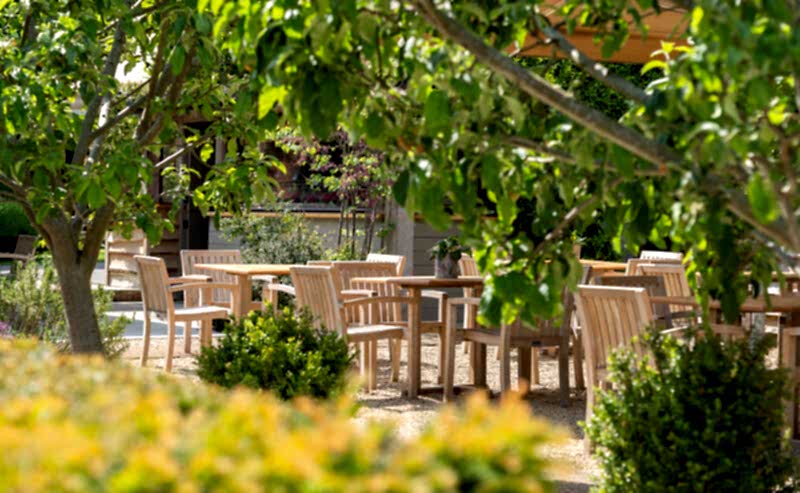 Hurley House - Wooden tables and chairs are arranged outdoors in a garden patio area, surrounded by green trees and shrubs on a sunny day.