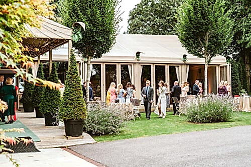 Hurley House - A group of people in formal attire gather outside a large white tent, surrounded by neatly trimmed trees and greenery, on a bright day. Some guests are mingling while others stand or sit at tables.