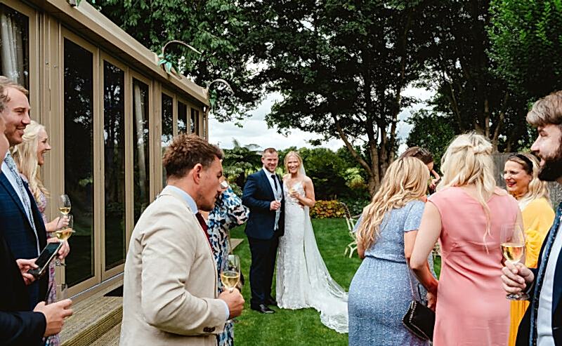 Hurley House - A bride and groom stand together smiling outside as wedding guests in colorful outfits gather around them, holding drinks and chatting on a grassy lawn near a building and trees.