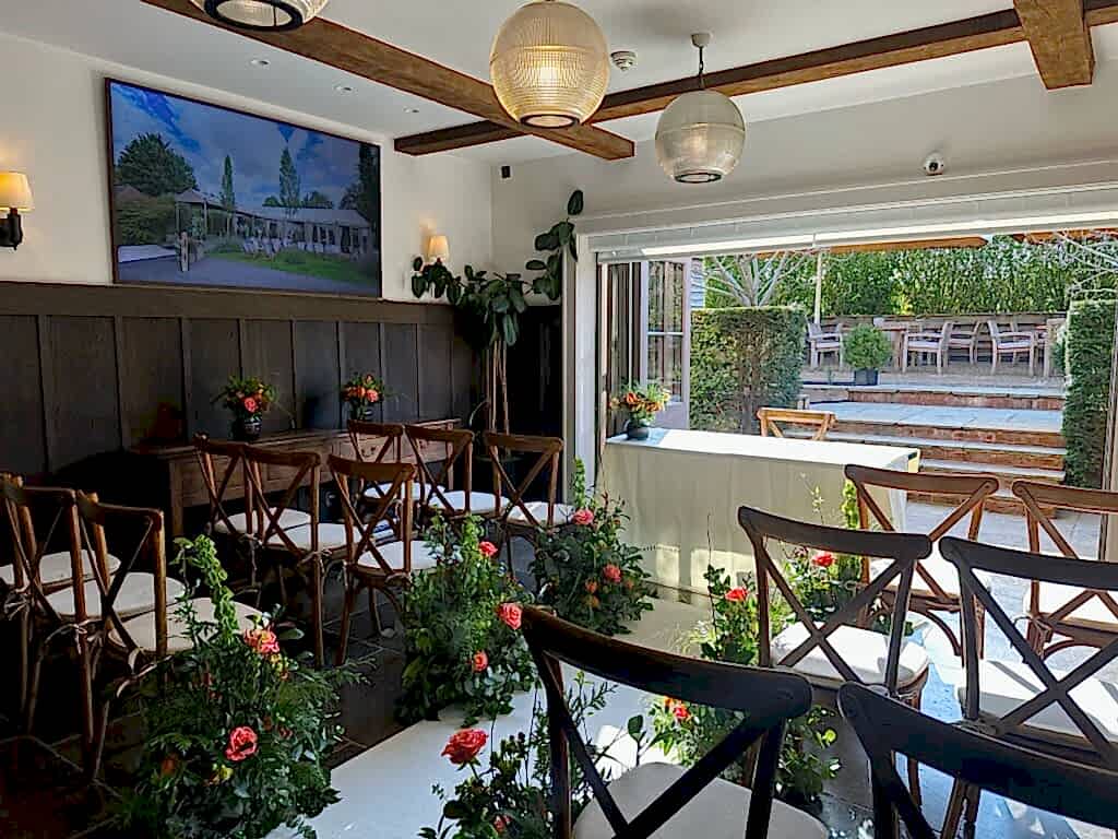Hurley House - A small, elegantly decorated room in a luxury wedding venue in Berkshire is set up for a ceremony. Wooden chairs are arranged in rows amidst lush floral arrangements. Large windows flood the space with natural light, and a photo adorns the wall. A table is gracefully positioned at the front.