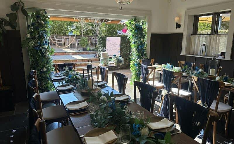 Hurley House - Elegant dining area with wooden tables and chairs, decorated with green foliage and blue accents. Outdoor view through an archway adorned with greenery, perfect for a luxury wedding venue in Berkshire. Bright, airy ambiance with paper lanterns hanging from the ceiling.