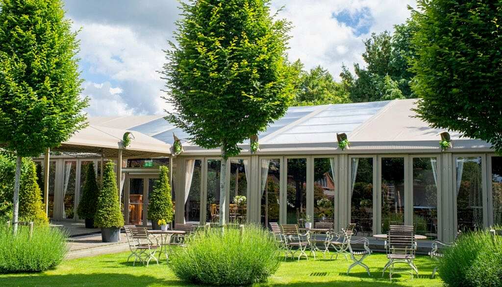 Hurley House - A luxury wedding venue in Berkshire features a charming outdoor dining area with tables and chairs set on lush green grass, surrounded by neatly trimmed bushes and tall trees. Large windows on the elegant building reflect the partly cloudy sky above.