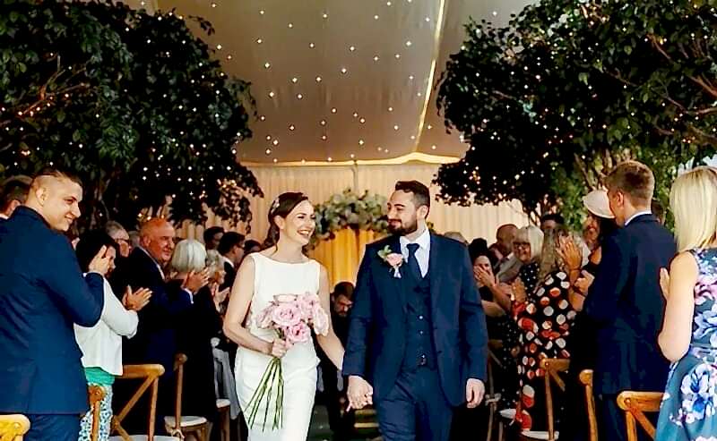 Hurley House - A couple walks down the aisle, holding hands. The bride wears a white dress and holds a bouquet, while the groom wears a dark suit. Guests on either side stand and applaud. The venue is decorated with trees and twinkling lights.