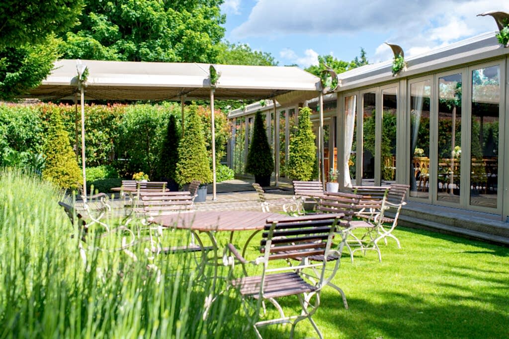Hurley House - An outdoor patio with empty metal tables and chairs on green grass, surrounded by lush trees and bushes. A canopy and glass-walled building are in the background under a blue sky with clouds.