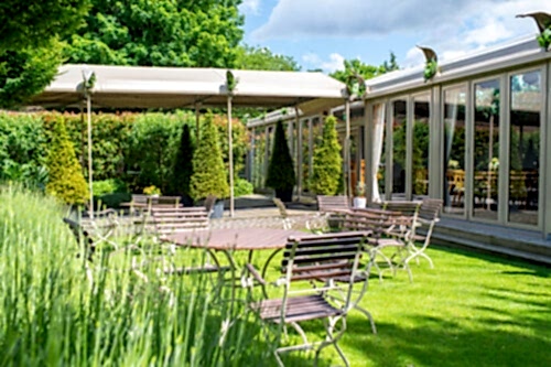 Hurley House - Outdoor seating area with metal tables and chairs on a green lawn, shaded by a canopy and surrounded by trimmed bushes and trees next to a building with large glass windows on a sunny day.