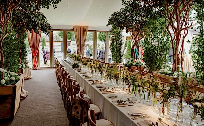 Hurley House - A long, elegant banquet table is set for a formal event, decorated with flowers, greenery, and candles under a canopy with string lights. Large windows and trees create a lush, garden-like atmosphere. People gather outside.