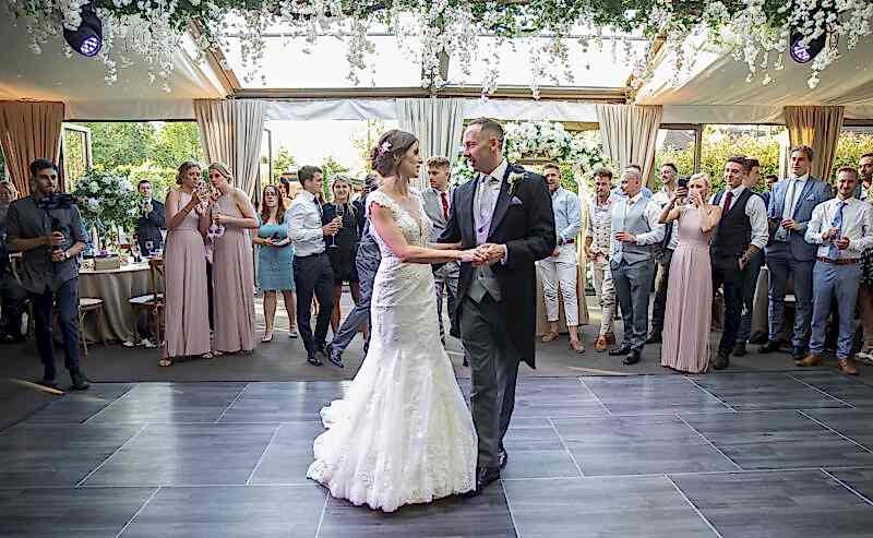 Hurley House - In a luxury wedding venue in Berkshire, the bride and groom share their first dance under soft lighting, surrounded by guests. She dazzles in a lace wedding dress, while he looks dapper in his suit. The room is adorned with elegant floral arrangements, creating an enchanting atmosphere.