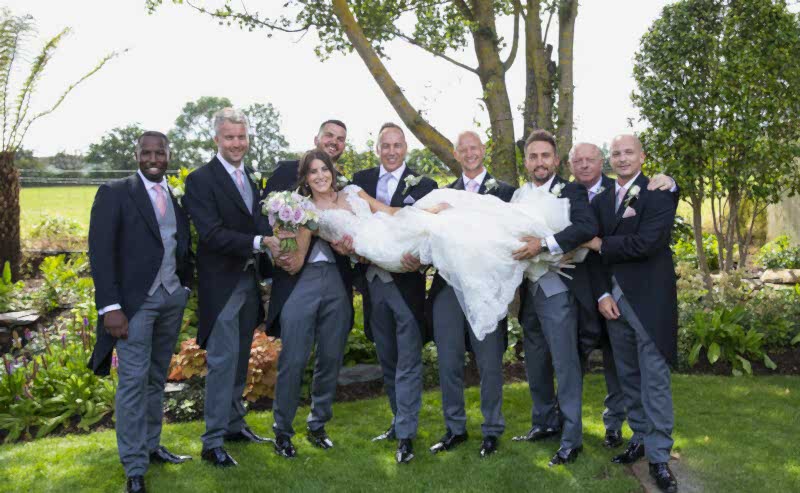 Hurley House - A bride in a lace wedding dress is playfully held horizontally by groomsmen in matching suits with pink ties, all smiling against the backdrop of a luxury wedding venue in Berkshire, surrounded by lush gardens and a sunny sky.