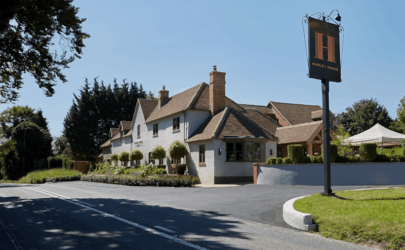 Hurley House - A well-maintained, two-story brick building with a pitched roof, surrounded by greenery on a sunny day. A large sign in front reads Hurley House. Theres a clear, blue sky above the building nestled by trees.