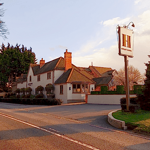 Hurley House - A quaint country hotel with a beige facade and brown roof sits beside a spacious parking area. The sign reads Hurley House Hotel. Tall trees border the property, and the sky is clear with soft sunlight illuminating the scene.