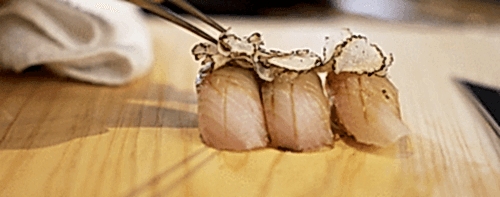 Hurley House - Three pieces of sushi topped with thinly sliced truffle are arranged on a wooden surface, with a hand using chopsticks to pick up one piece.