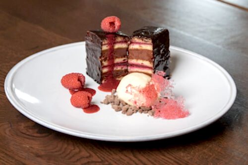 Hurley House - A plated dessert featuring layered chocolate-covered cake with fruit filling, garnished with raspberries, a scoop of vanilla ice cream, chocolate crumbles, and pink sugar tuile on a white plate.