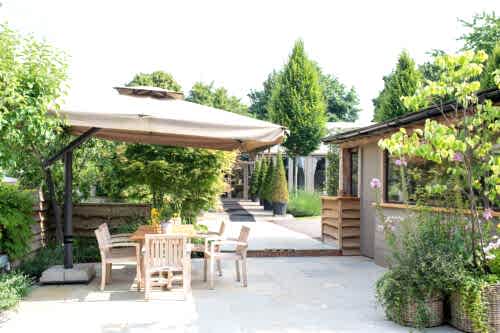 Hurley House - A sunny patio with a wooden table and four chairs under a large beige umbrella, surrounded by green plants and trees, near a house with large windows and potted plants.