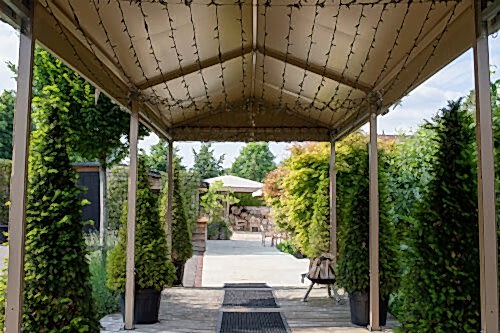 Hurley House - A covered walkway with string lights overhead leads to a sunny patio area with tables, chairs, and lush green plants on both sides. Tall potted bushes line the walkway, and trees and shrubs surround the patio.