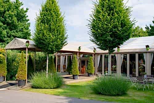 Hurley House - A garden venue with tall manicured trees, large windows, and a covered walkway leading to an entrance. Outdoor seating and lush greenery surround the building under a partly cloudy sky.