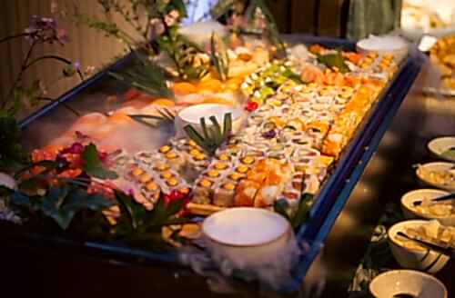 Hurley House - A large platter of assorted sushi rolls and nigiri, garnished with green leaves and flowers, is displayed on a bed of ice at a buffet in a luxury wedding venue in Berkshire, with several small bowls of dipping sauces nearby.
