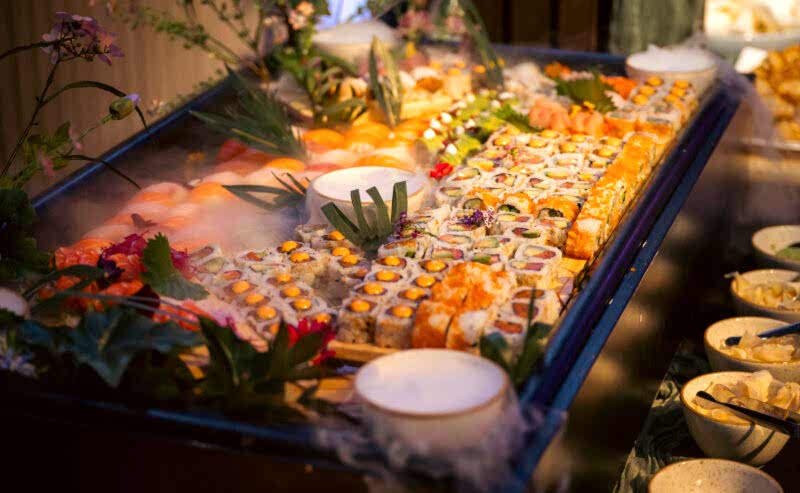 Hurley House - A large platter of assorted sushi rolls and nigiri, garnished with green leaves and flowers, is displayed on a bed of ice at a buffet in a luxury wedding venue in Berkshire, with several small bowls of dipping sauces nearby.