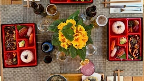 Hurley House - A top-down view of a table set with Japanese bento boxes featuring rice, grilled fish, sashimi, and vegetables. Yellow flowers are arranged in the center, with drinks and chopsticks also visible.