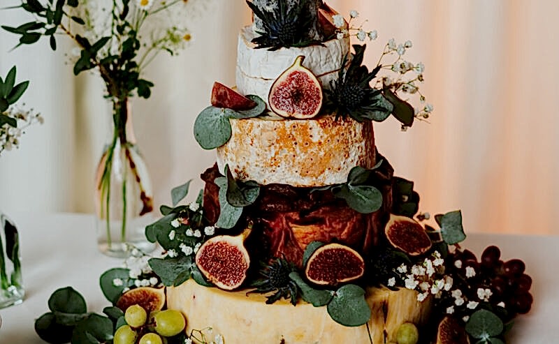 Hurley House - A three-tiered cheese cake is adorned with figs, grapes, and eucalyptus leaves, epitomizing the elegance of a luxury wedding venue in Berkshire. Encircled by small boxes of crackers on a white tablecloth, it sits alongside vases filled with greenery and white flowers.