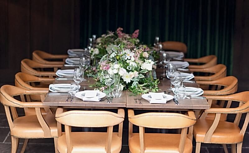 Hurley House - A long dining table is elegantly set with white plates, folded napkins, and glassware at a boutique wedding venue in Berkshire. The centerpiece features lush floral arrangements with green and pink tones. Surrounding the table are ten tan leather chairs.