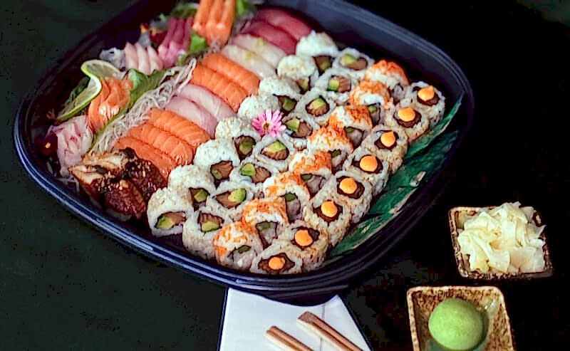 Hurley House - A platter of assorted sushi and sashimi with rolls, nigiri, and garnishes on a green tablecloth. Chopsticks, wasabi, pickled ginger, and two menus are set beside the tray. A small floral arrangement is in the background.
