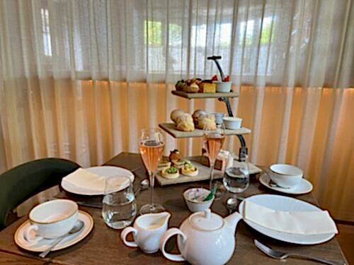 Hurley House - A table set for afternoon tea with two place settings, a white teapot, glasses of pink sparkling wine, and a three-tiered stand with assorted pastries, scones, and sandwiches. Light filters through sheer curtains behind.