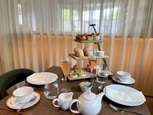 Hurley House - A table set for afternoon tea with two glasses of pink sparkling drink, teapots, cups, plates, and a three-tiered stand holding pastries, scones, and sandwiches, against a sheer curtain backdrop.
