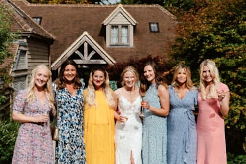 Hurley House - Seven women stand close together, smiling outdoors in front of a house. They wear colorful dresses, and one in the center is in white bridal attire, suggesting a wedding celebration. Sunlight and greenery surround them.